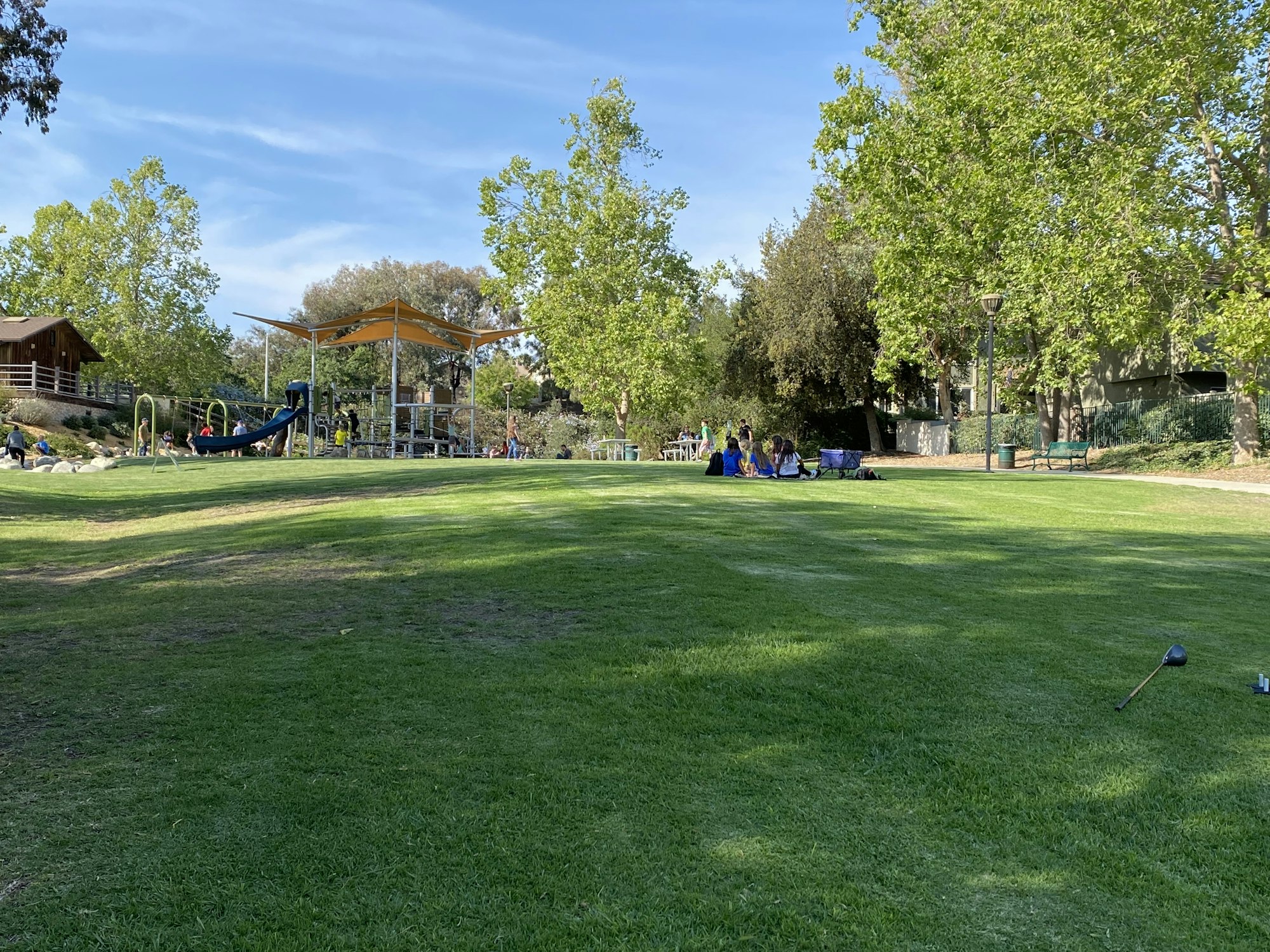 A park with a playground, green grass, trees, and people gathered around in groups.