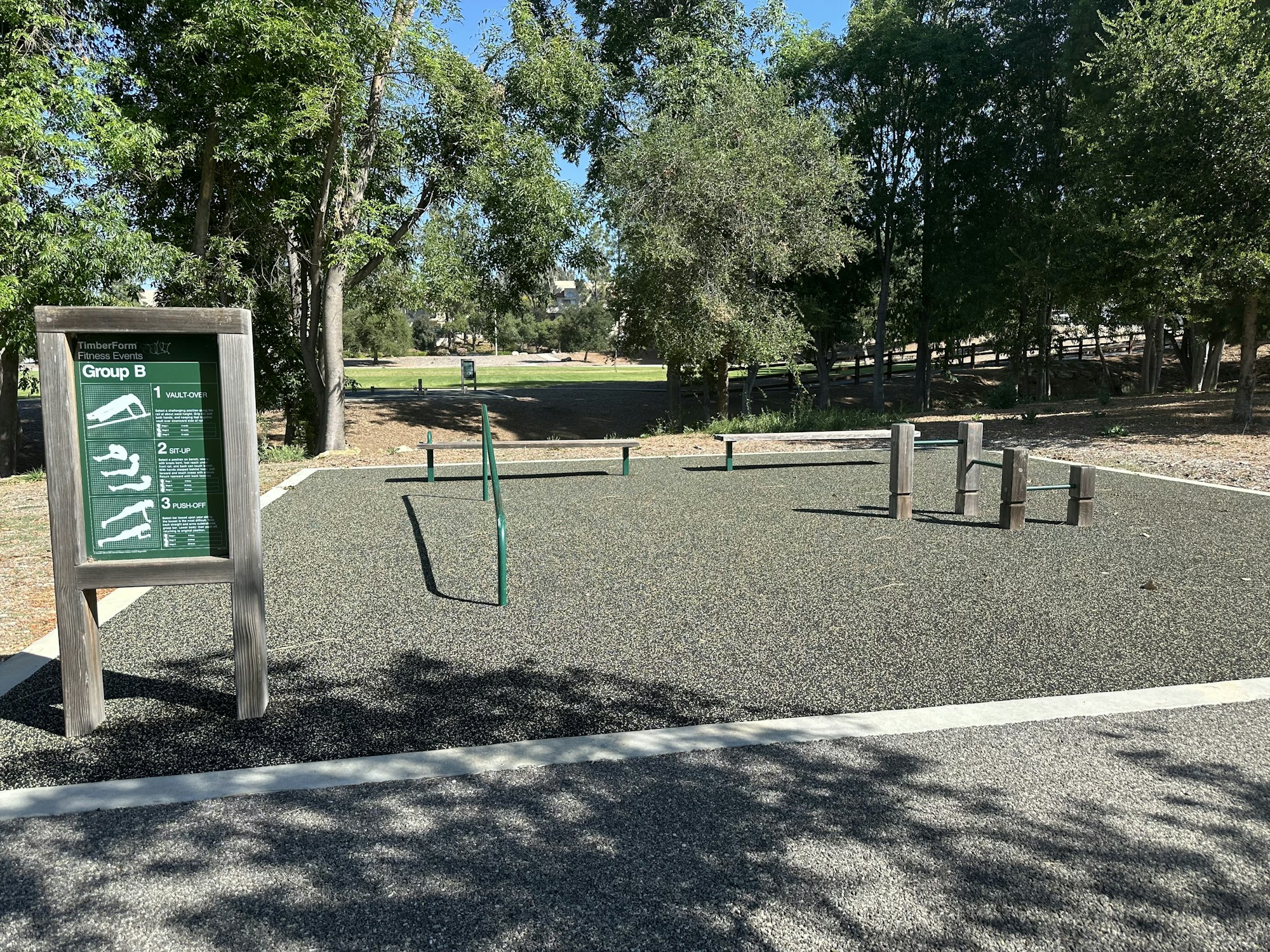 An outdoor fitness area featuring equipment for exercises like vault over, step-up, and push-off, surrounded by trees.