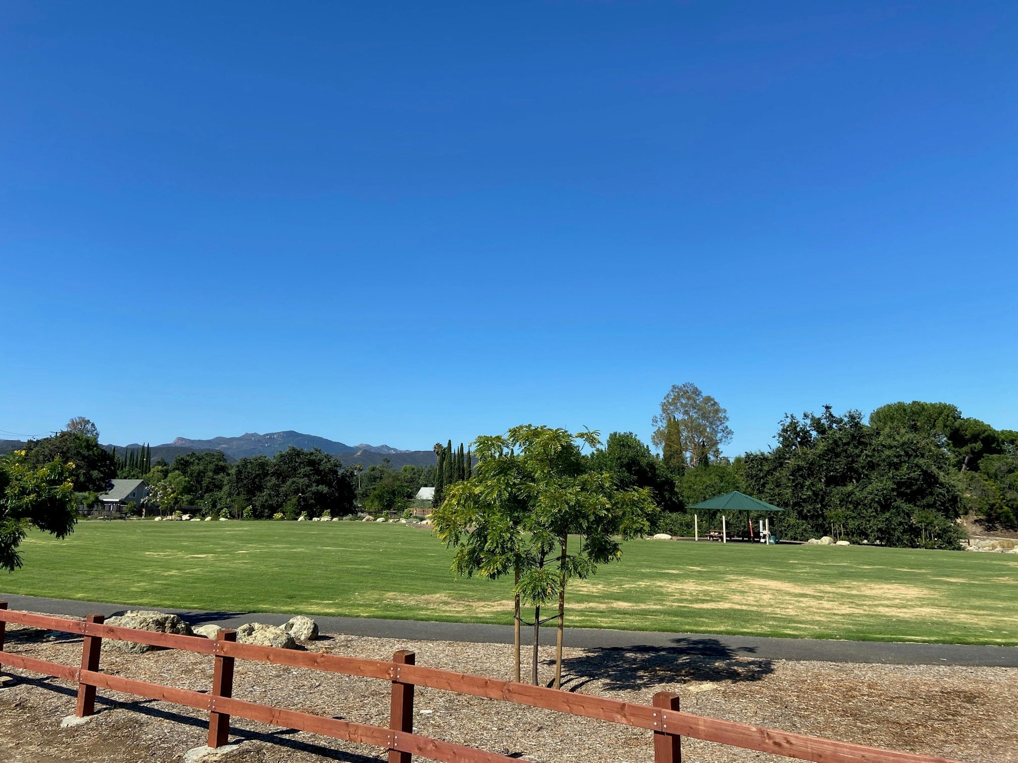 Open grassy area with trees, a gazebo, and a mountain in the background under a clear blue sky.