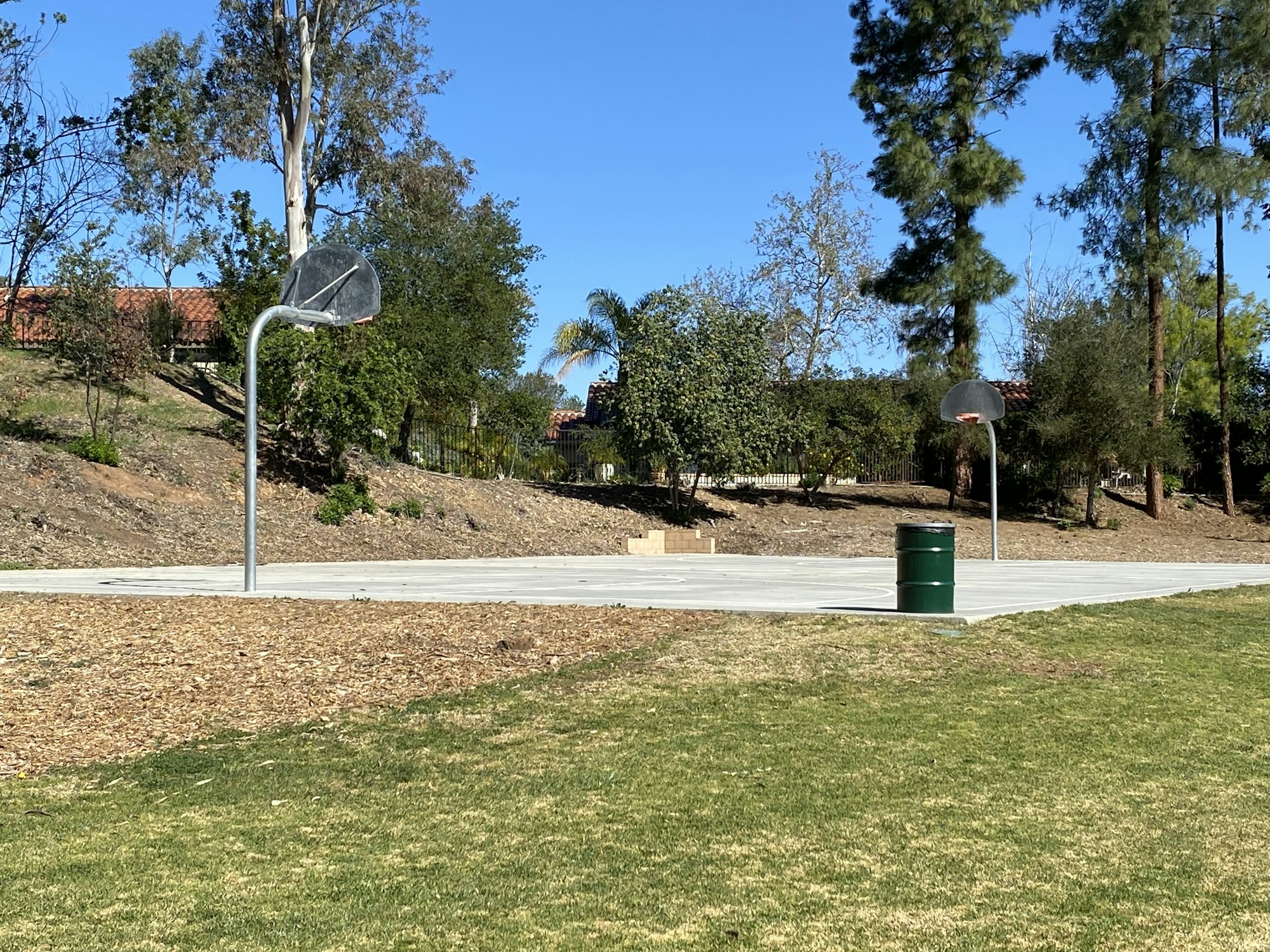 Two outdoor basketball hoops, a trash can, and trees in a park setting under a clear blue sky.