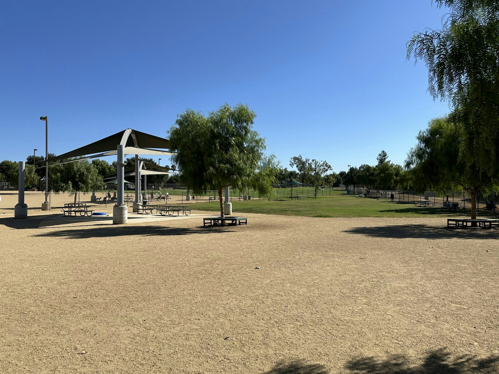 A park with picnic tables, shade structures, dirt field, grassy area, and trees under a clear blue sky.