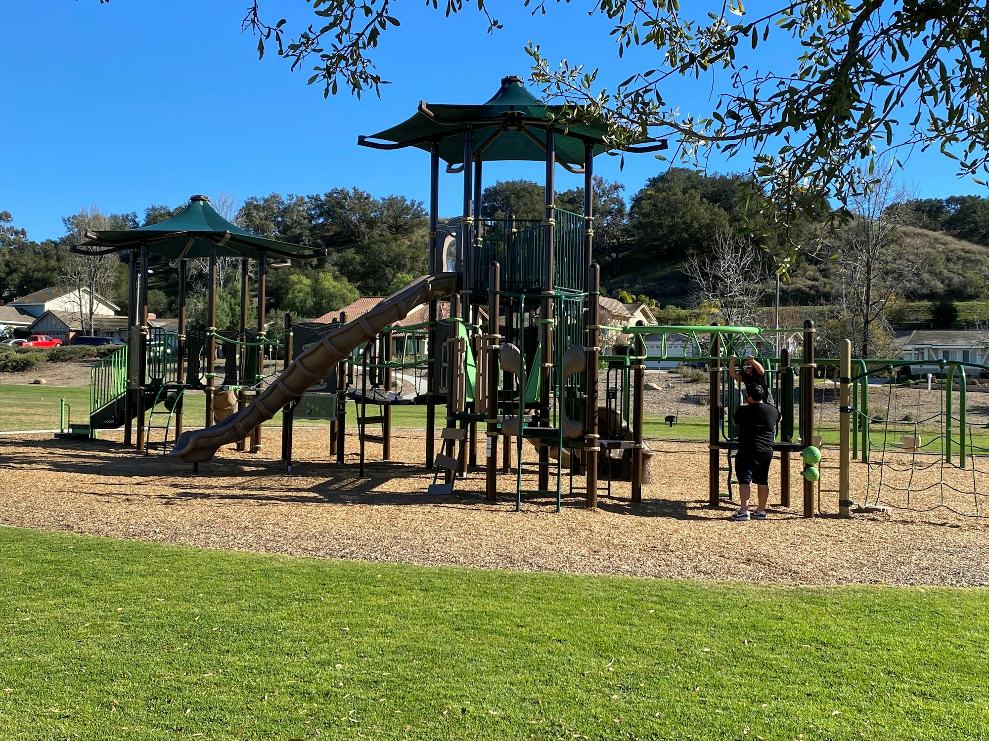 A playground with slides and climbing structures, surrounded by grass, with a person standing nearby.