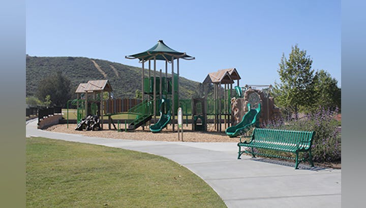 A playground with slides and climbing structures, surrounded by grass and trees, and a green bench nearby on a paved path.