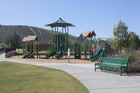 A playground with slides and climbing structures, surrounded by grass and trees, and a green bench nearby on a paved path.