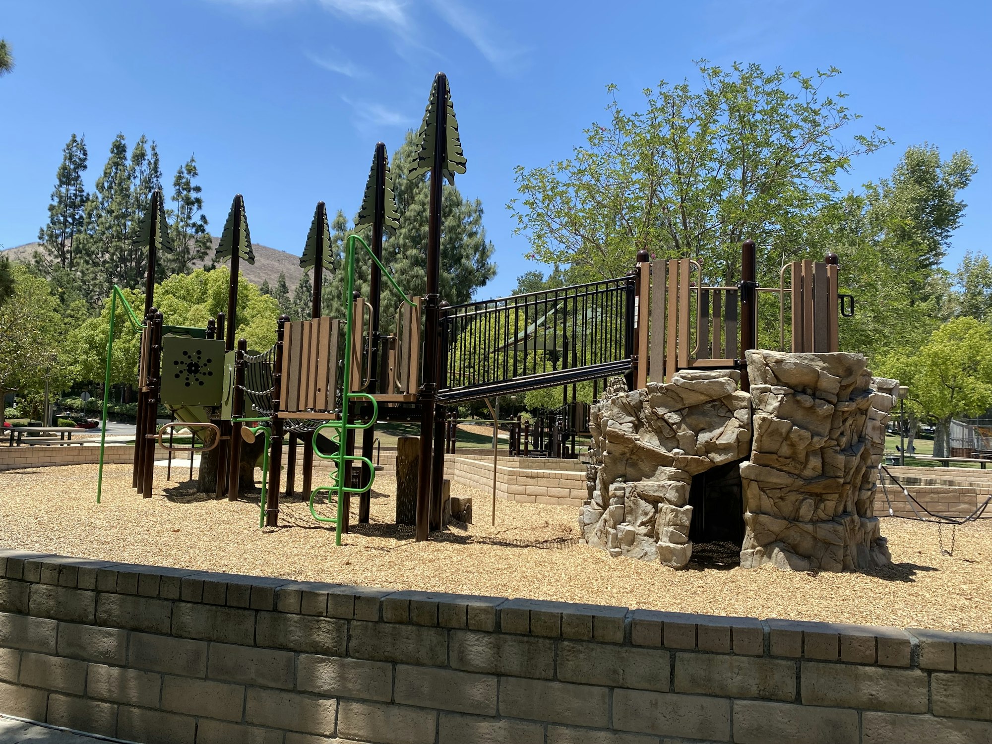 Playground with climbing structures, a bridge, and faux rock formations, surrounded by trees on a sunny day.