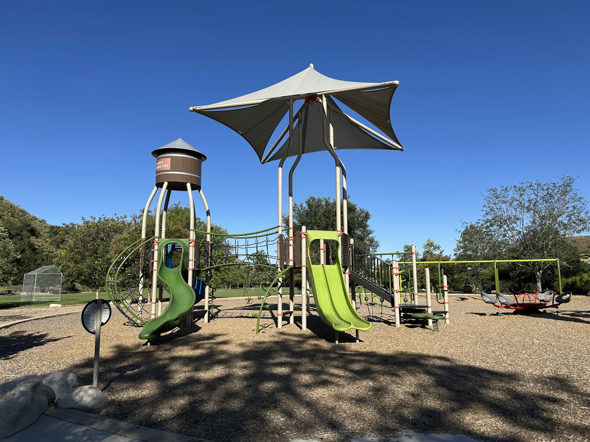 A sunny playground featuring green slides, climbing structures, swings, and a shaded area, surrounded by trees and blue skies.