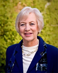 A person with short blonde hair wearing a blue blazer and necklace, standing outdoors with greenery in the background.