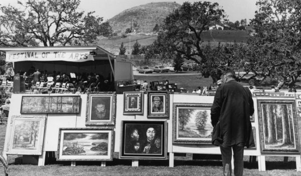 Outdoor art festival with paintings displayed and a man viewing them, trees and hills in the background.