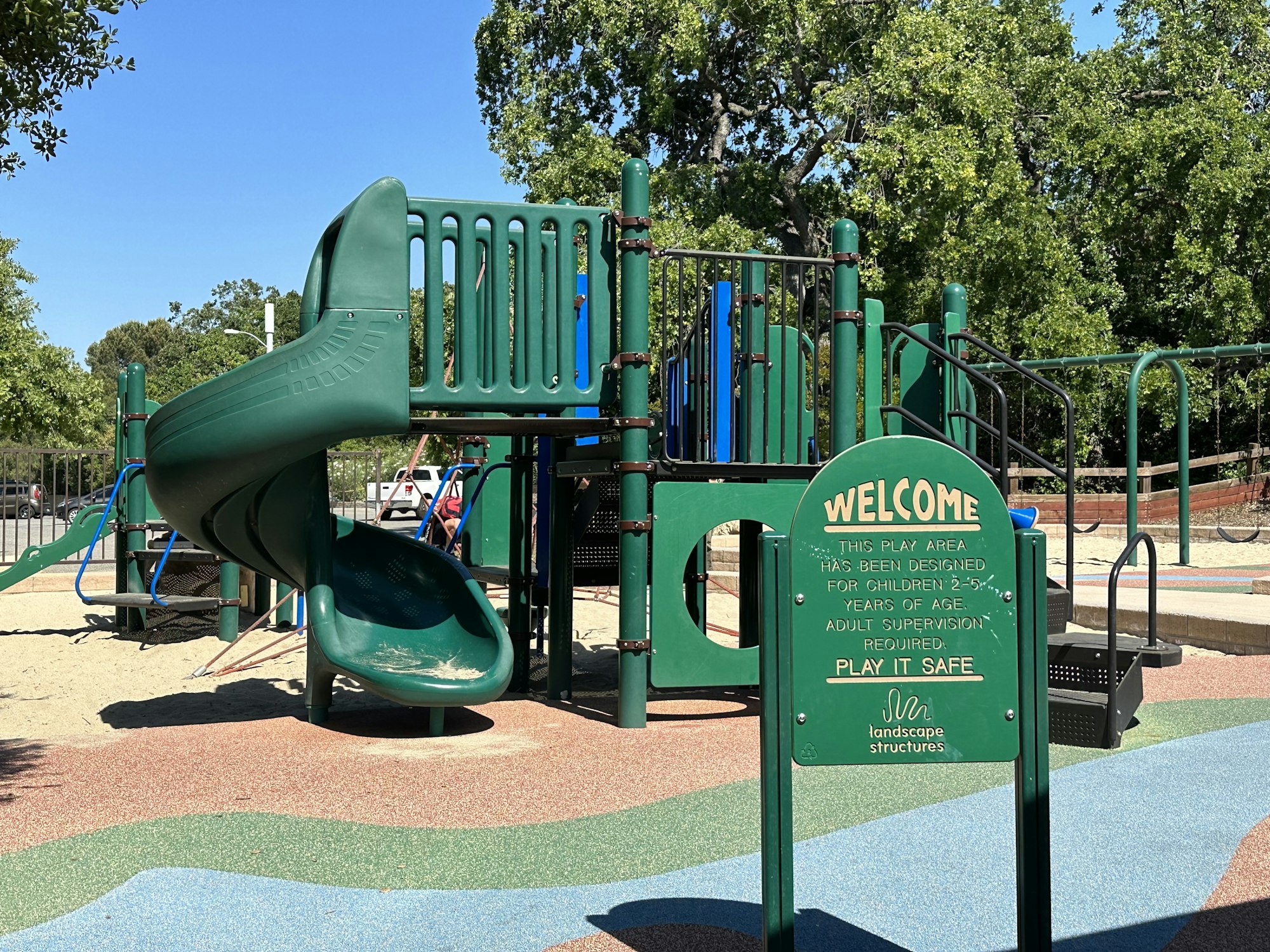 Playground with green slide, climbing equipment, and a sign welcoming children ages 2-5 with adult supervision required.