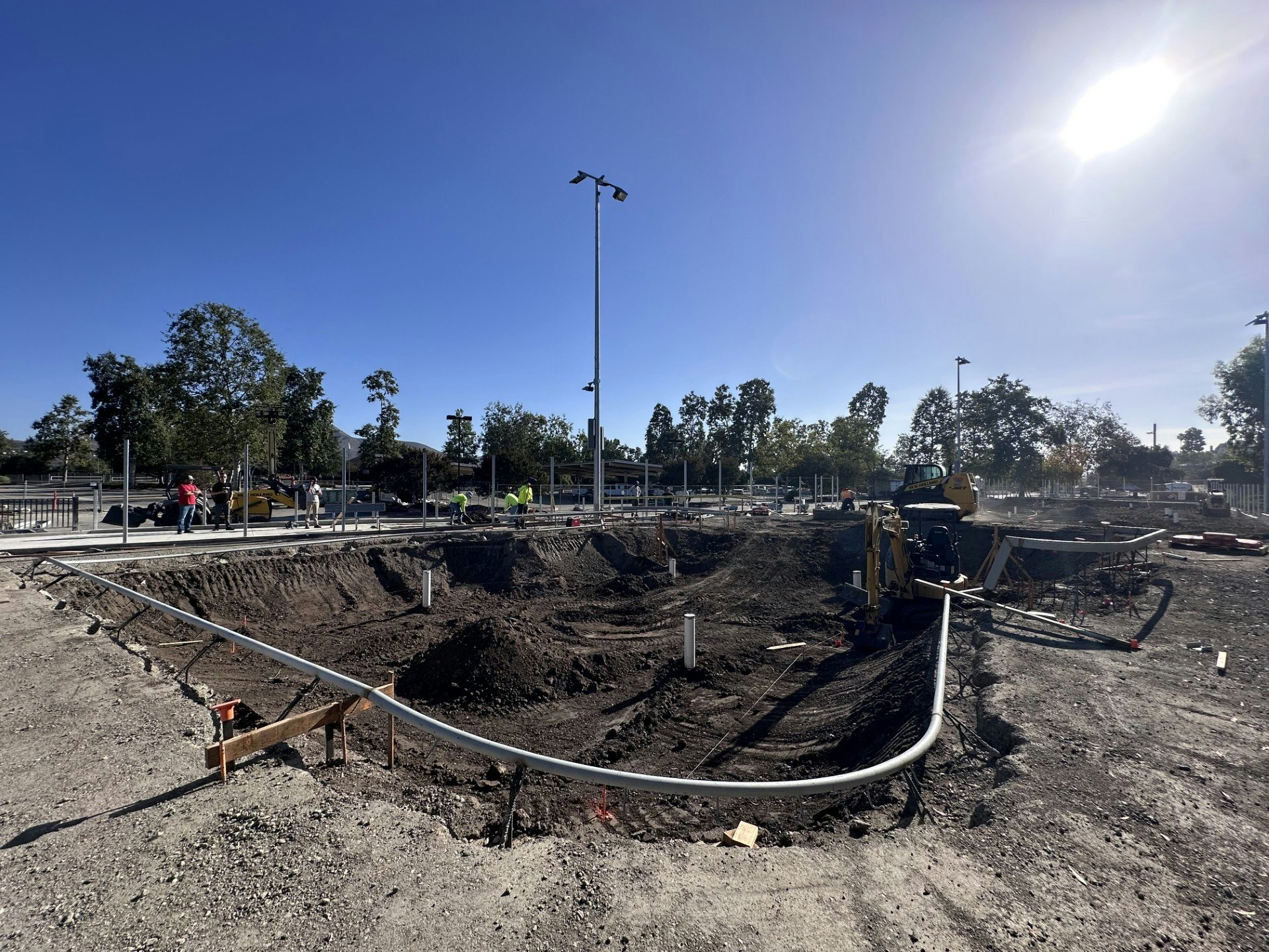 The image shows a construction site with workers, machinery, and trenches being dug, under a bright blue sky.