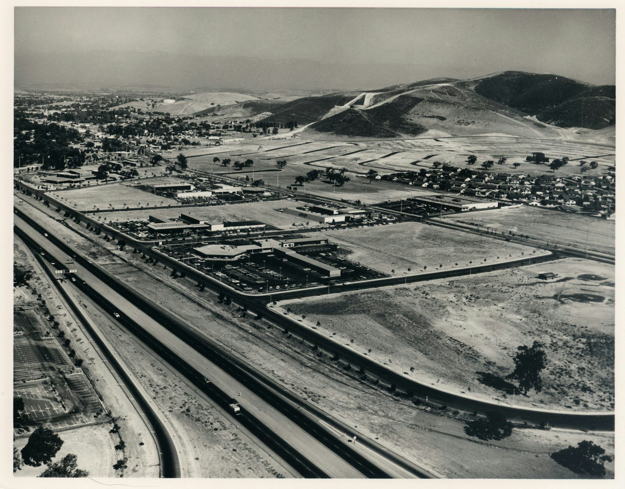 Aerial view of a mid-20th-century suburban area with roads, buildings, and hills in the background.