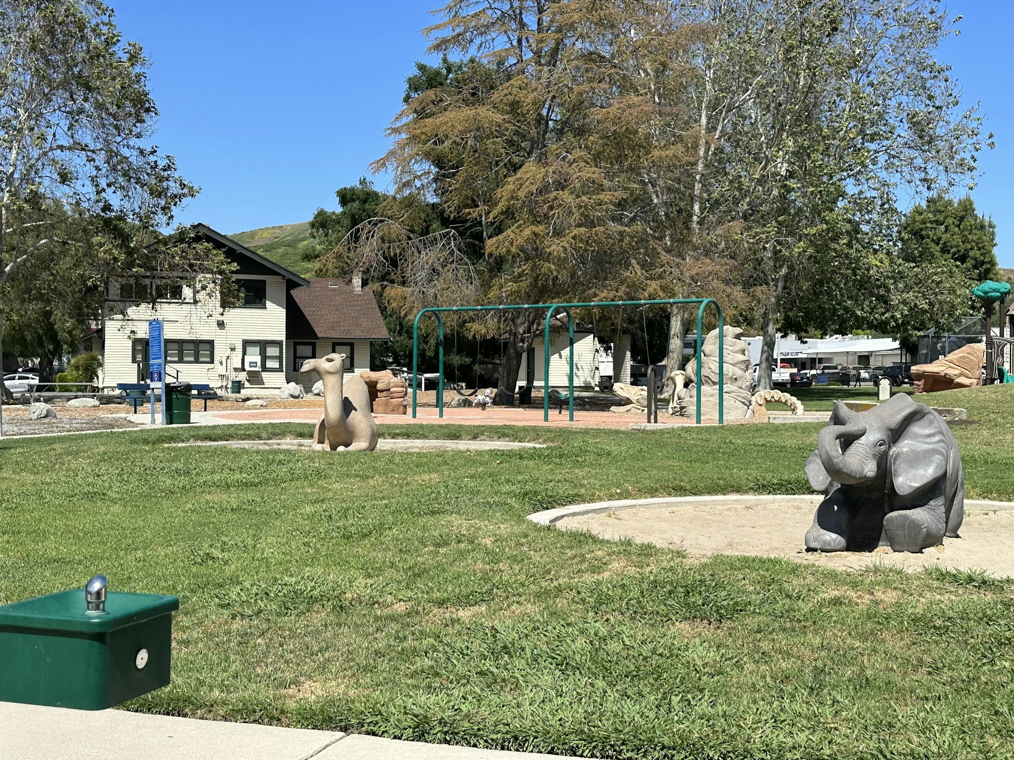A park with animal sculptures (elephant, camel), grass, trees, play area, swings, and a house in the background under a clear blue sky.