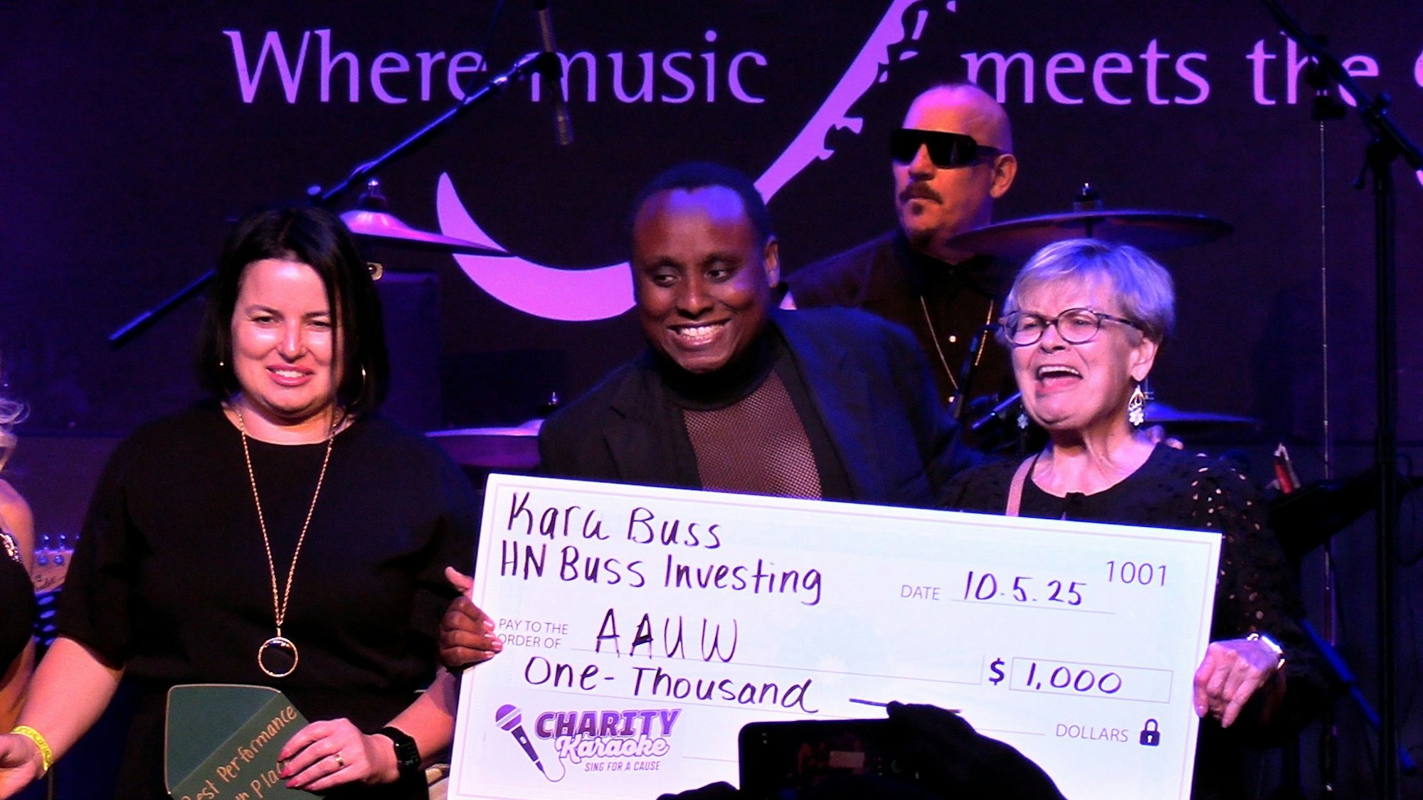 Three people holding a large charity check for $1,000 at a music-themed event.