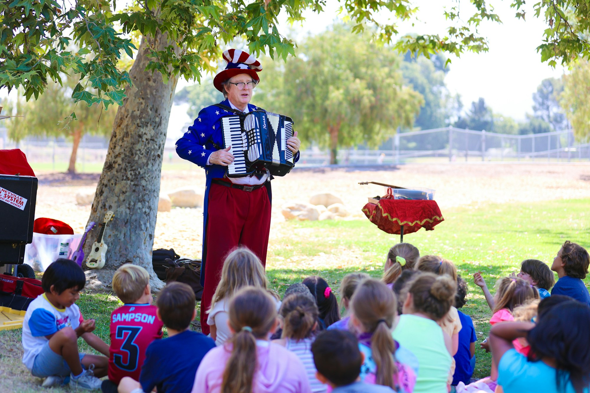 A performer in a starry costume plays an accordion under a tree for a group of children seated on the grass.