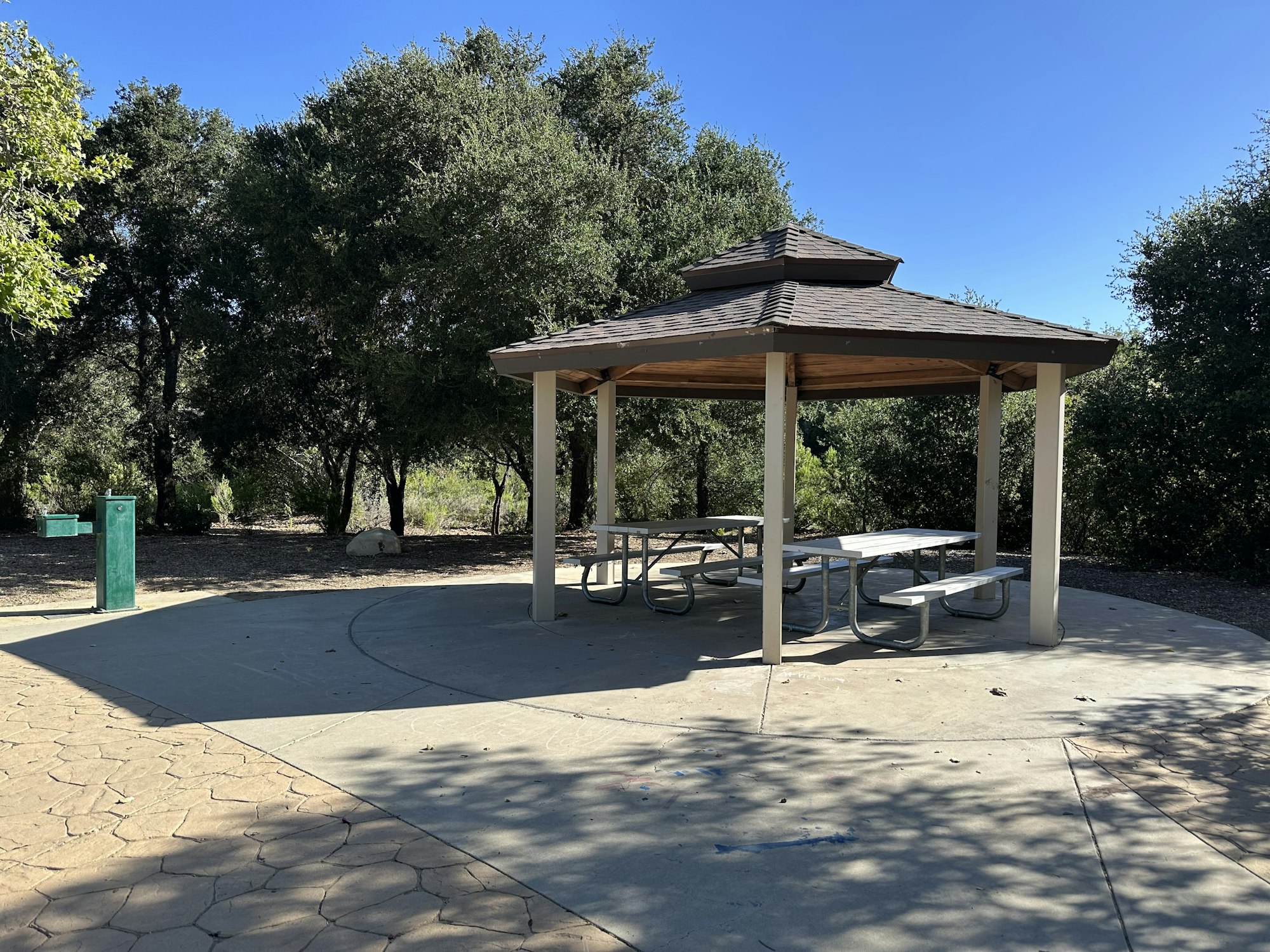 A gazebo with picnic tables surrounded by trees on a sunny day, with a nearby water fountain visible.