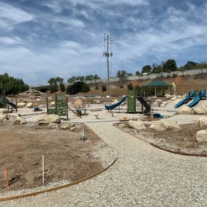 A playground with slides, climbing structures, and a path surrounded by rocks and sparse vegetation under a partly cloudy sky.