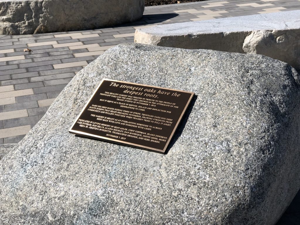A plaque on a rock in a paved area reads, "The strongest oaks have the deepest roots," dedicated to various individuals, dated November 1, 2019.