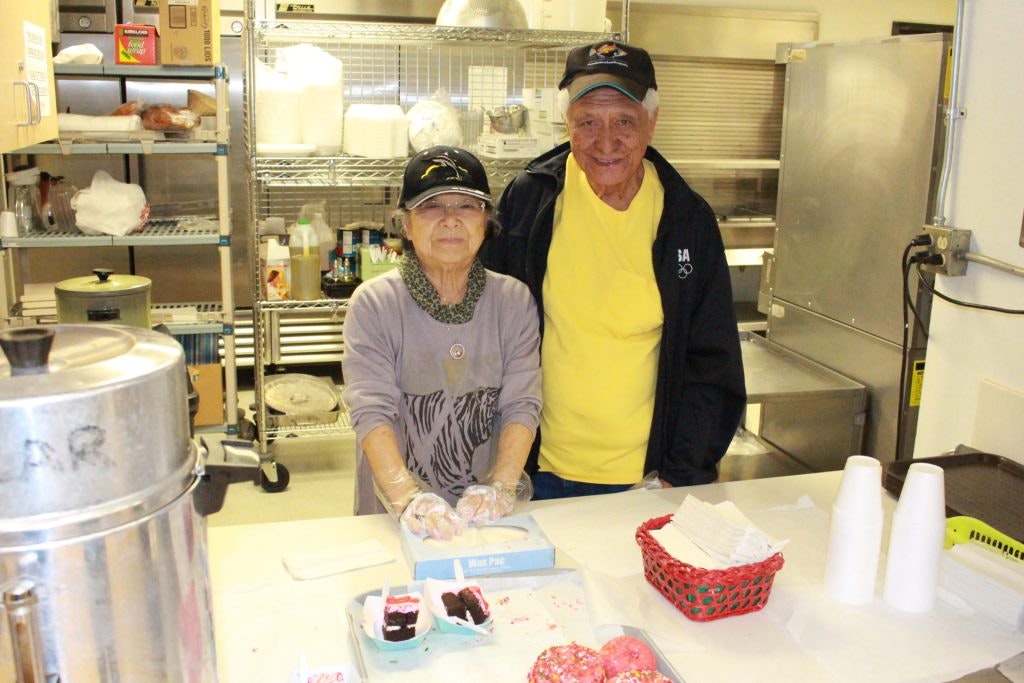 Two people in a kitchen setting, with baked goods and a drink dispenser on the table.