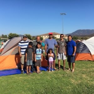 A group of people, including children, stand smiling in front of tents on a grassy field with a building and mountains in the background.