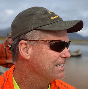 Man in a cap and sunglasses outdoors near water; another person blurred in the background.