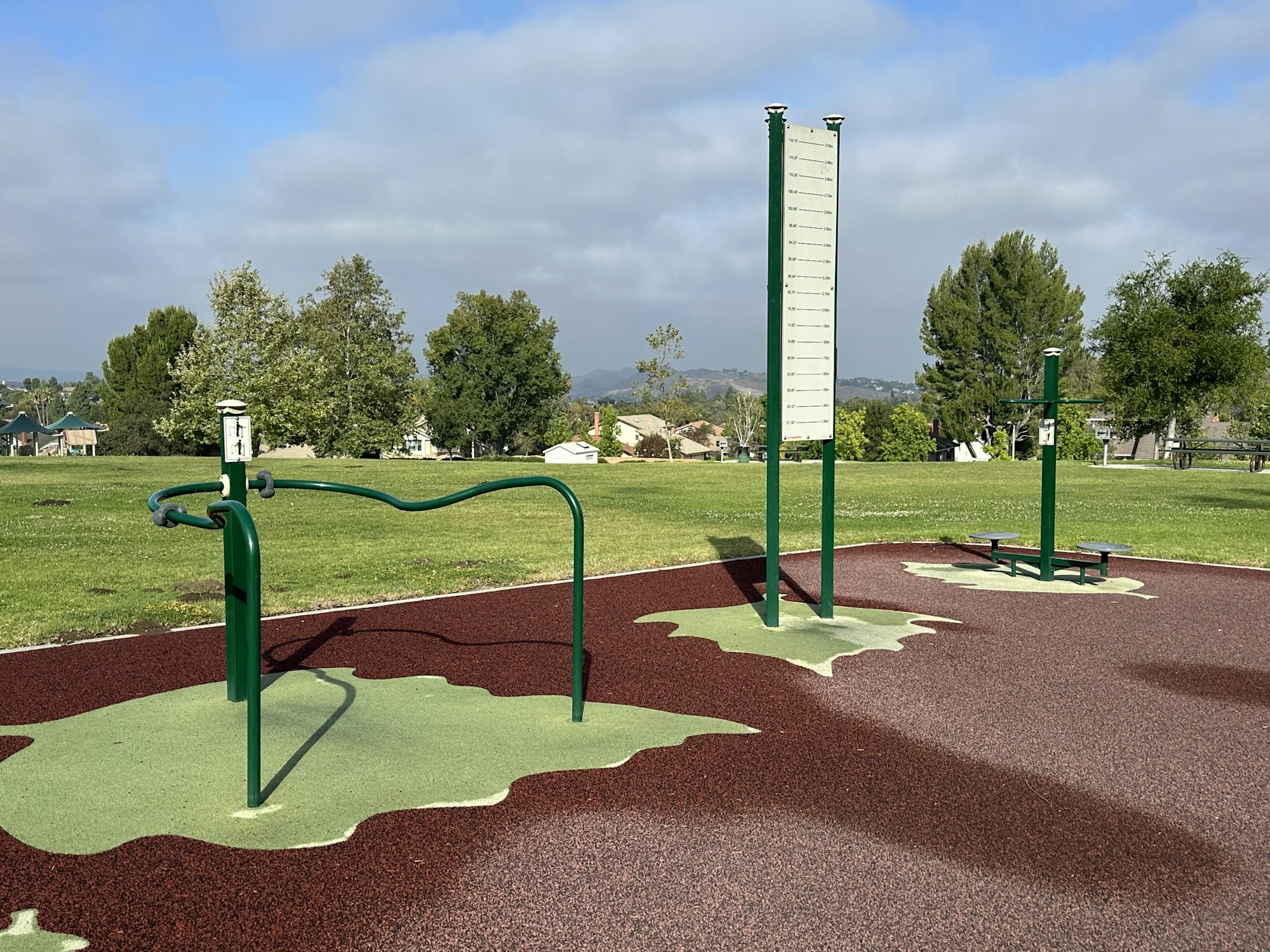 An outdoor fitness area with exercise equipment on colorful rubber flooring, surrounded by grass and trees under a cloudy sky.