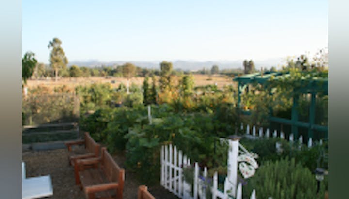 A garden with wooden benches, a white picket fence, lush plants, a trellis, and a scenic countryside backdrop.
