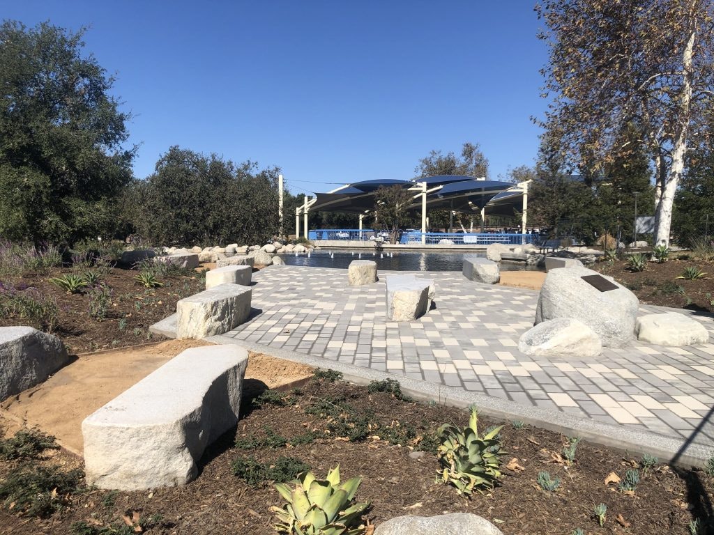 A landscaped park area with stone benches, patterned paving, trees, and a shaded structure.