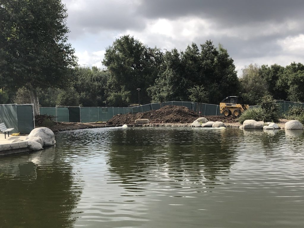 A pond with rocks and a construction area, featuring a small yellow bulldozer, surrounded by trees and a cloudy sky.