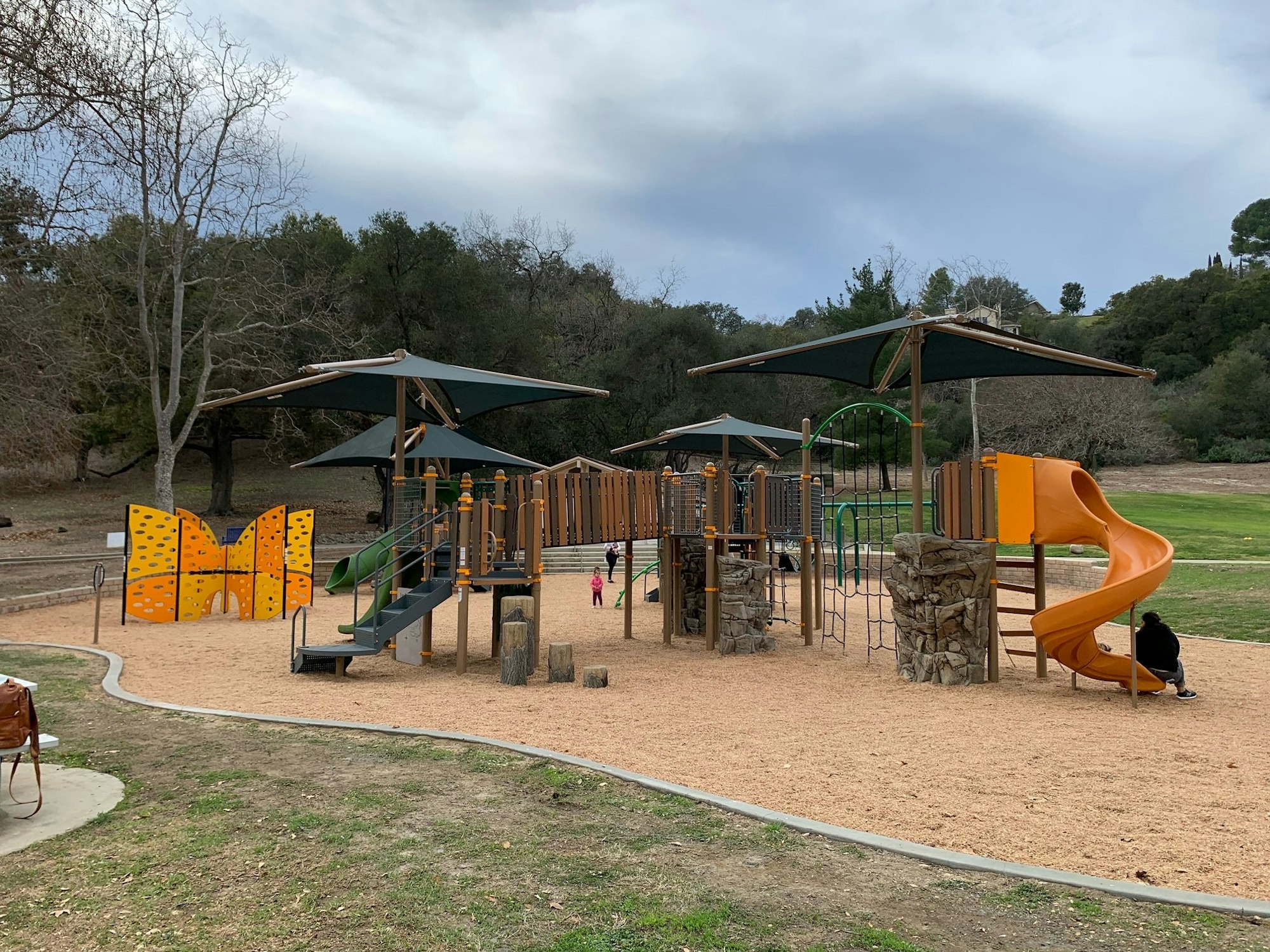A playground with slides, climbing structures, and shade canopies, surrounded by trees and grass.