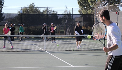 People playing tennis on a court, some holding rackets, with others in the background.