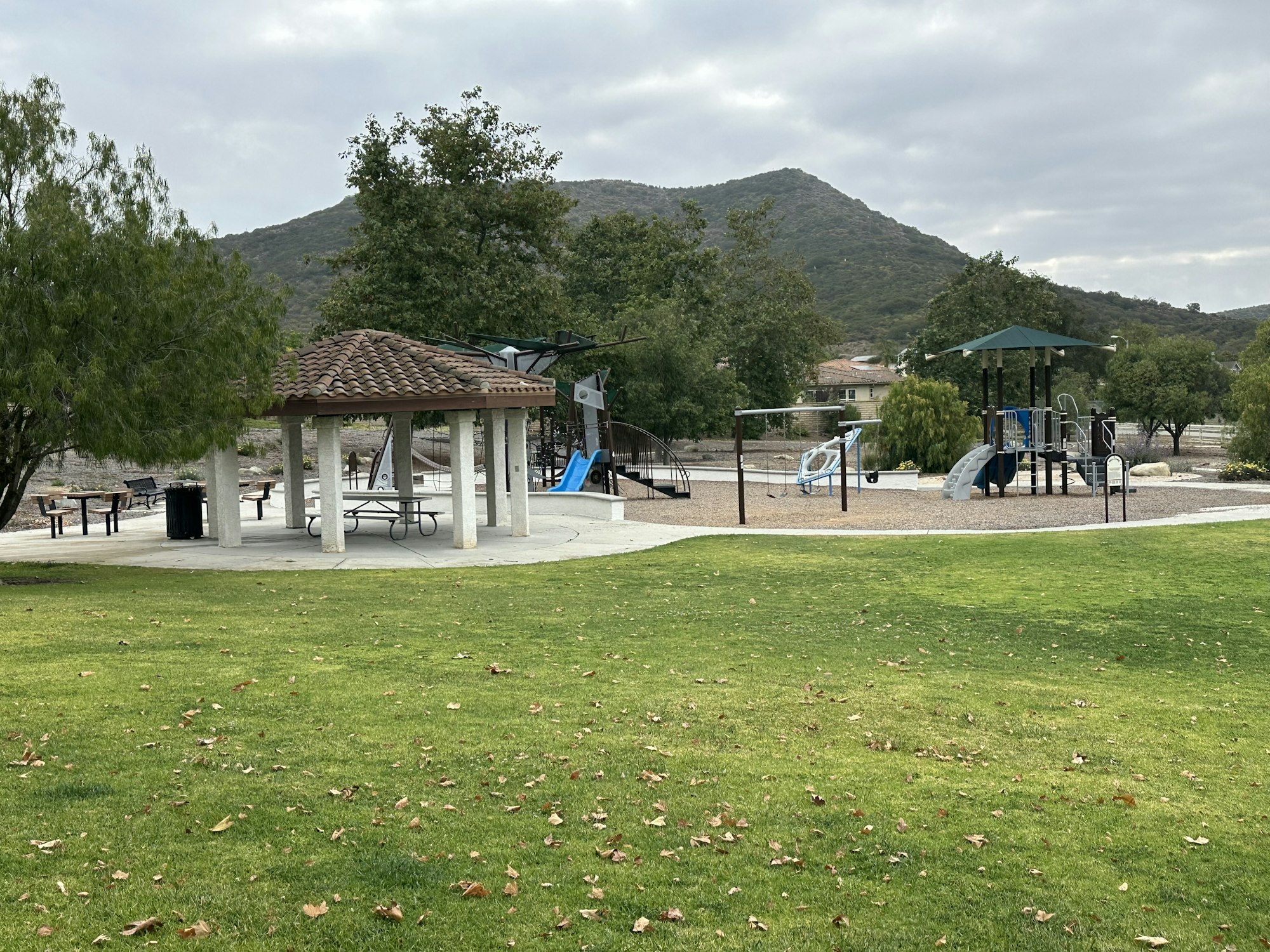 A park scene featuring a covered picnic area, playground equipment, and a grassy field with mountains in the background.