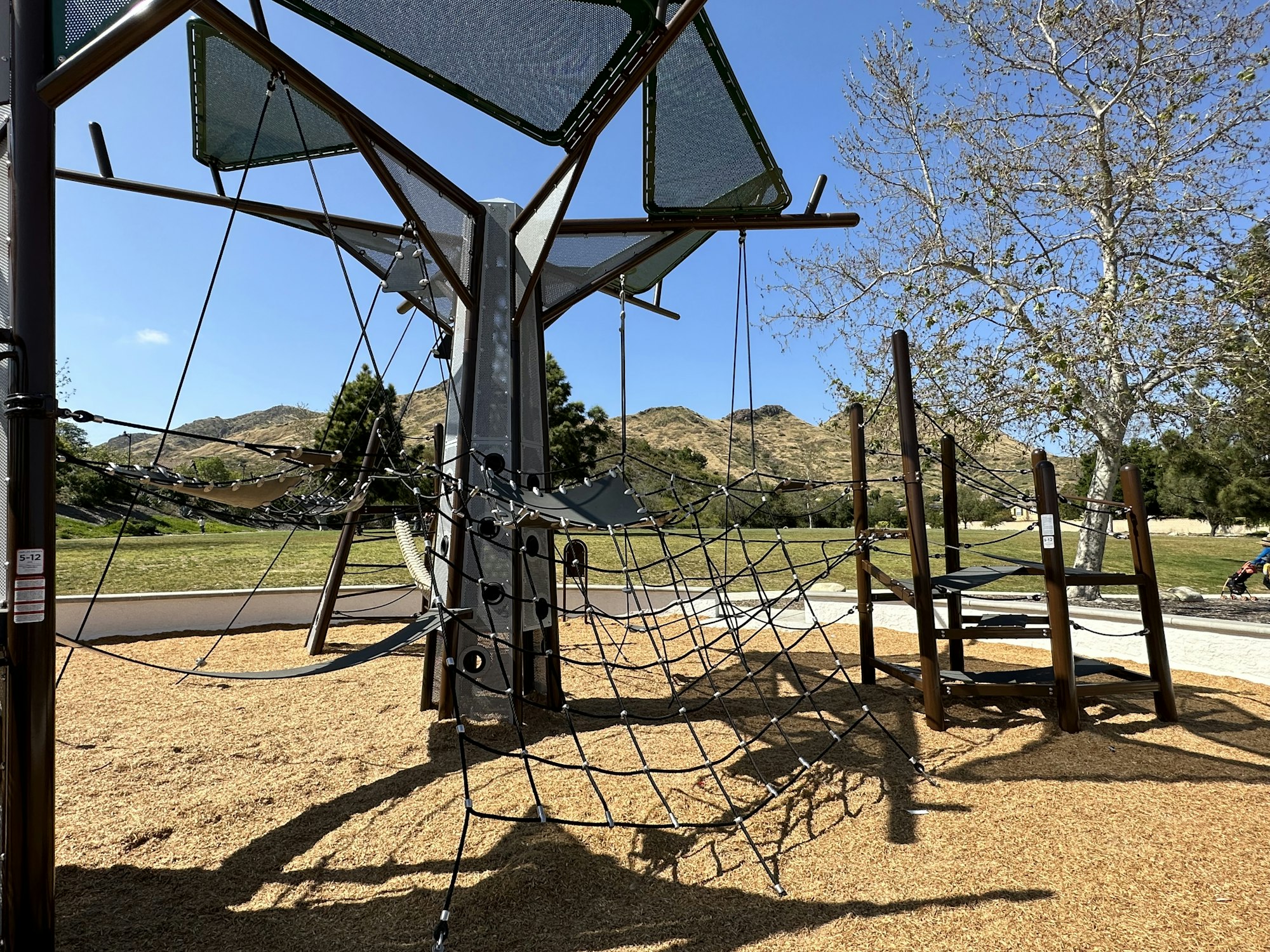 Modern playground with climbing nets and structures, set in a park with trees and hills in the background.