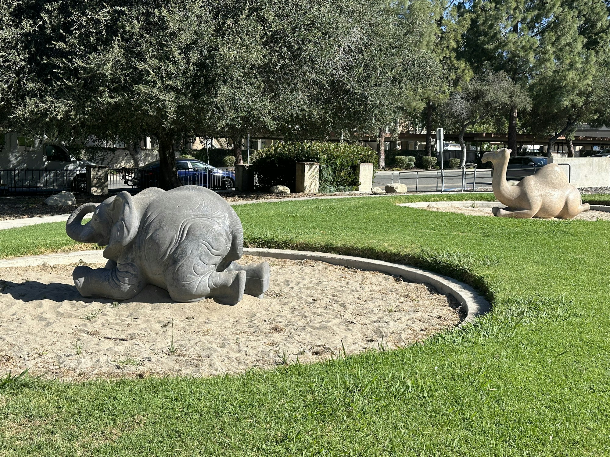 The image shows two animal sculptures: a grey elephant on the left and a beige camel on the right, both in a grassy area.