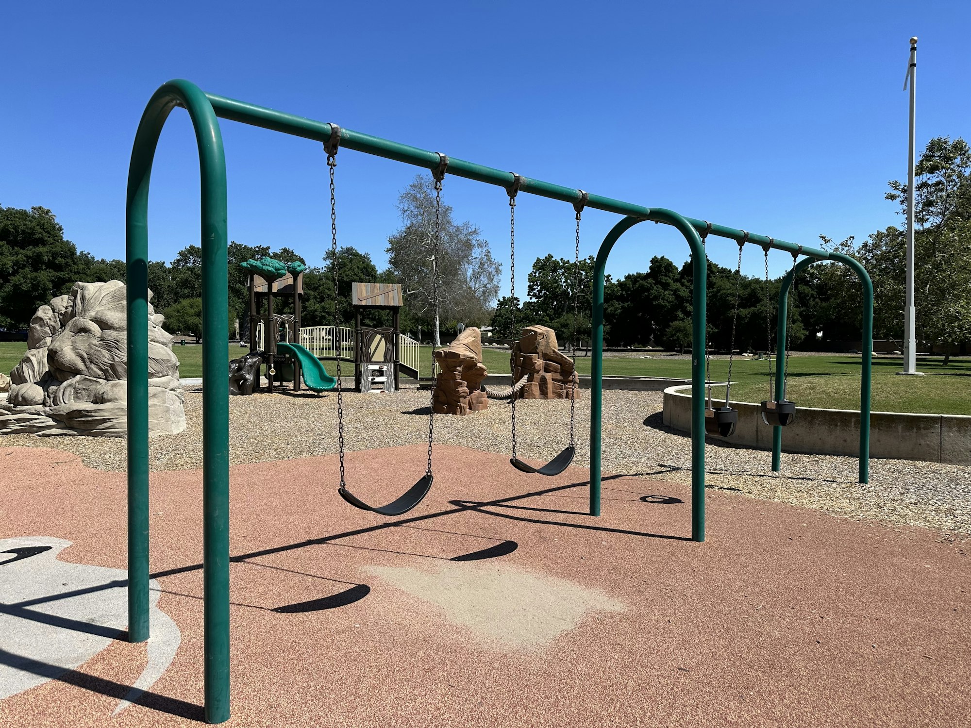 A playground swing set with four seats, a climbing structure, and slides in the background under a clear blue sky.