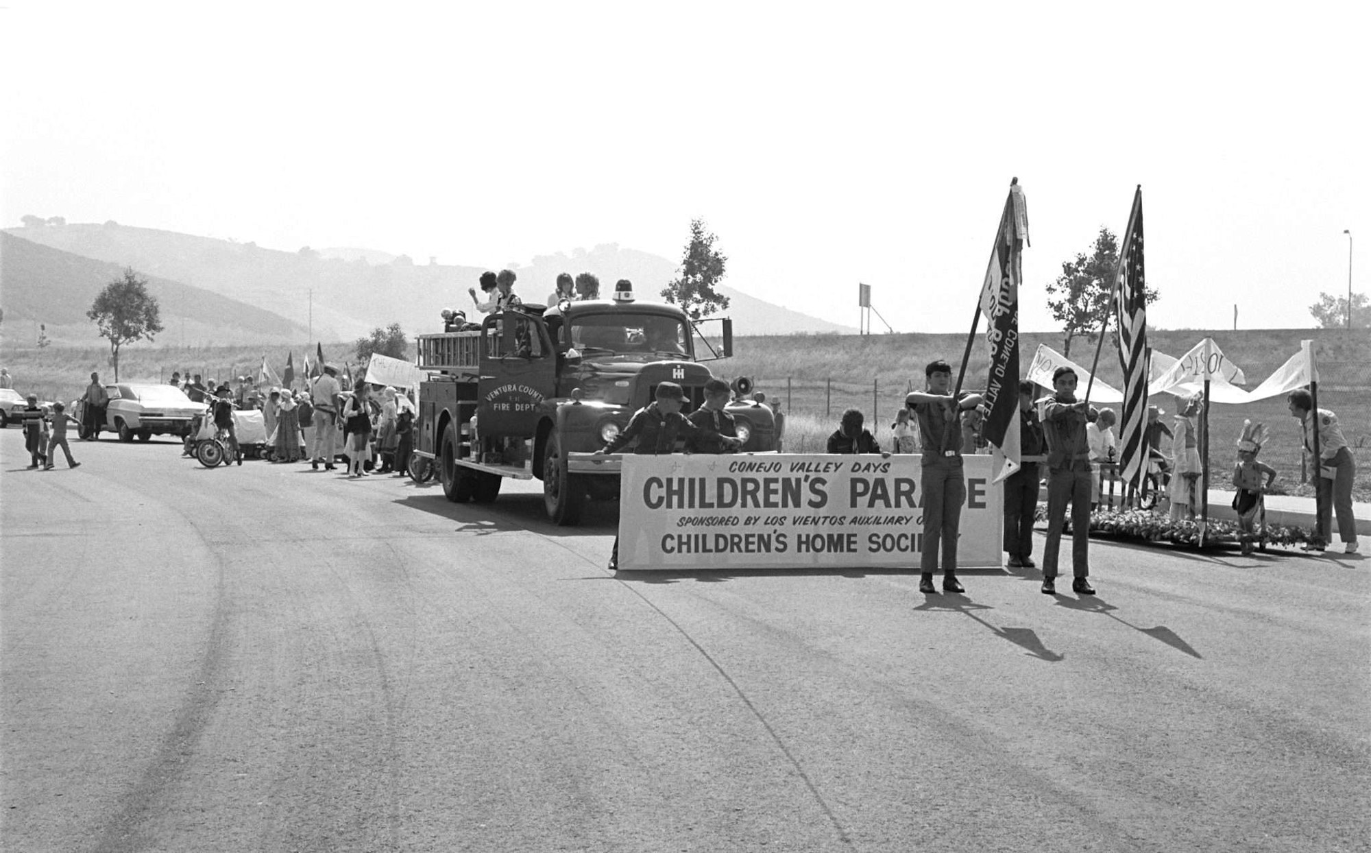 A parade with a fire truck, people holding signs and flags for Conejo Valley Days Children's Parade on a road.