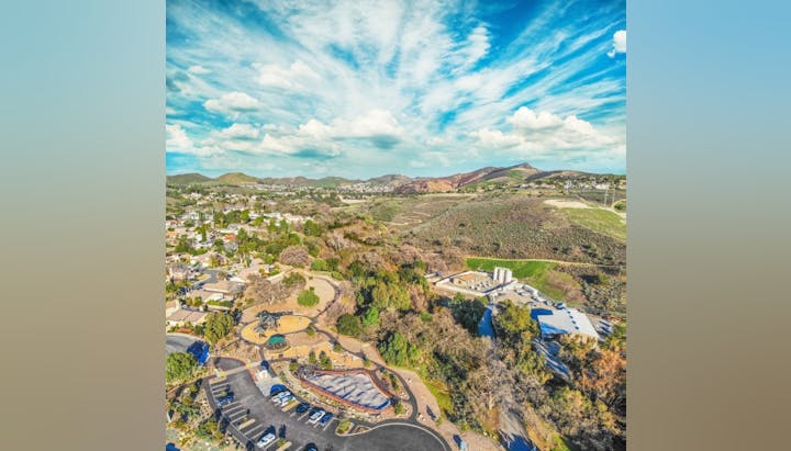 Aerial view of a suburban area with houses, a park, parking lot, greenery, and hills under a blue sky with clouds.