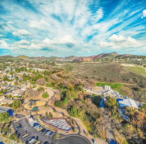 Aerial view of a suburban area with houses, a park, parking lot, greenery, and hills under a blue sky with clouds.