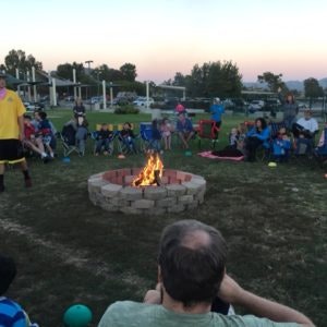 People sitting in a circle around a small campfire in a park setting during the evening.