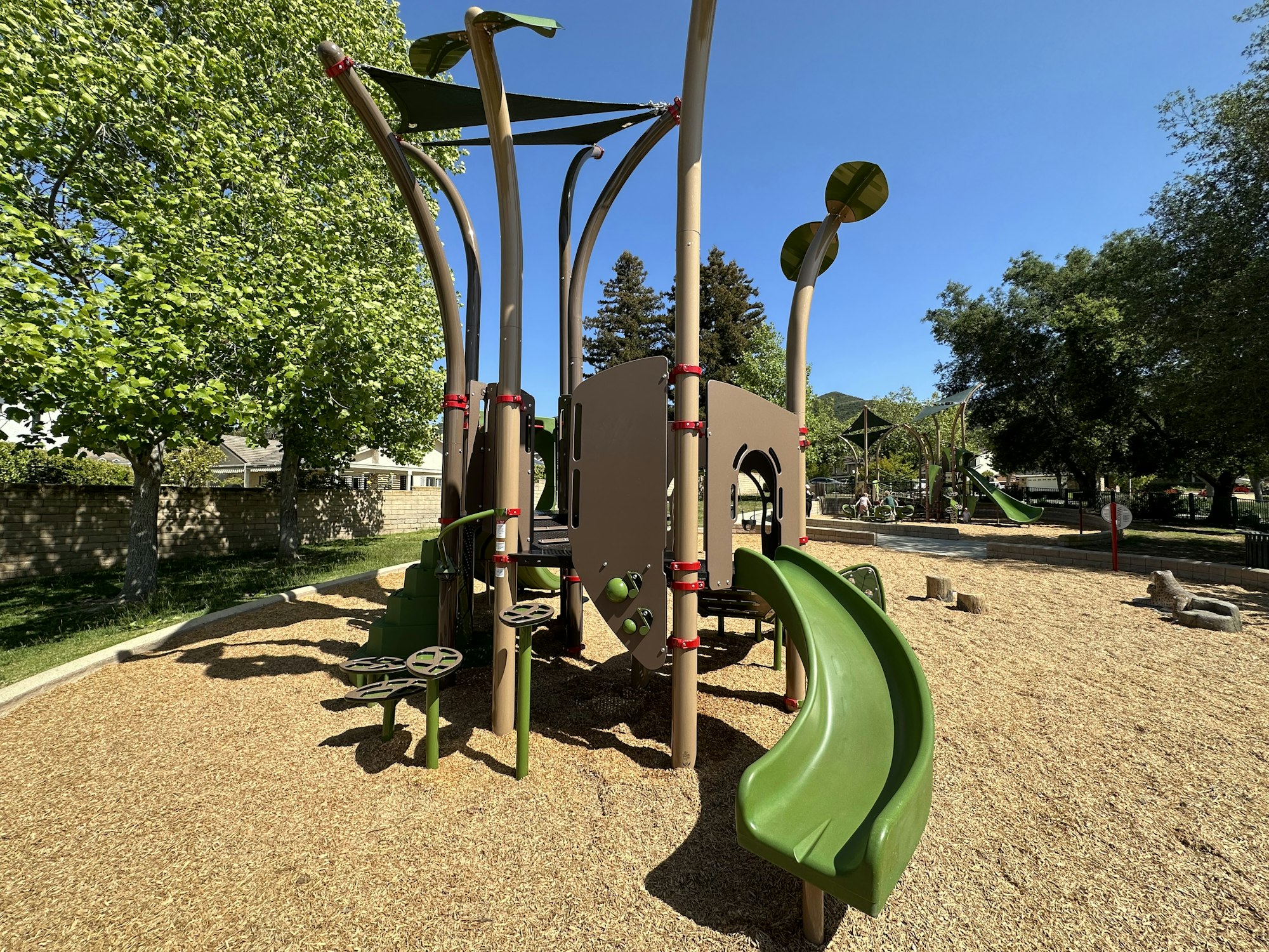 A playground with slides, climbing structures, surrounded by trees on a sunny day.