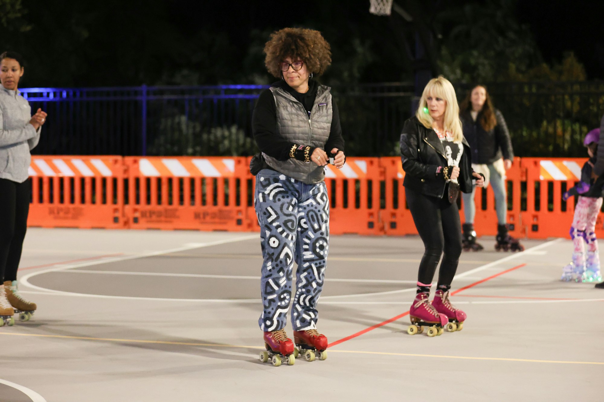 The image shows a roller skating event at night with several people skating and a court surrounded by orange barriers.
