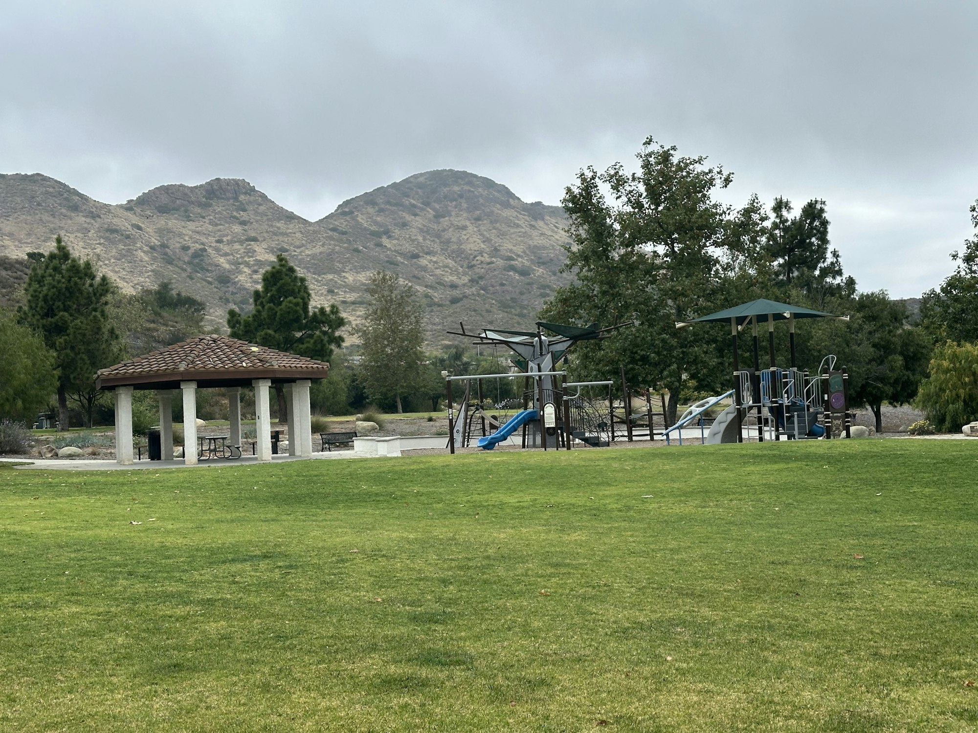 A park scene featuring a gazebo, playground equipment, and green grass, with mountains in the background and overcast skies.