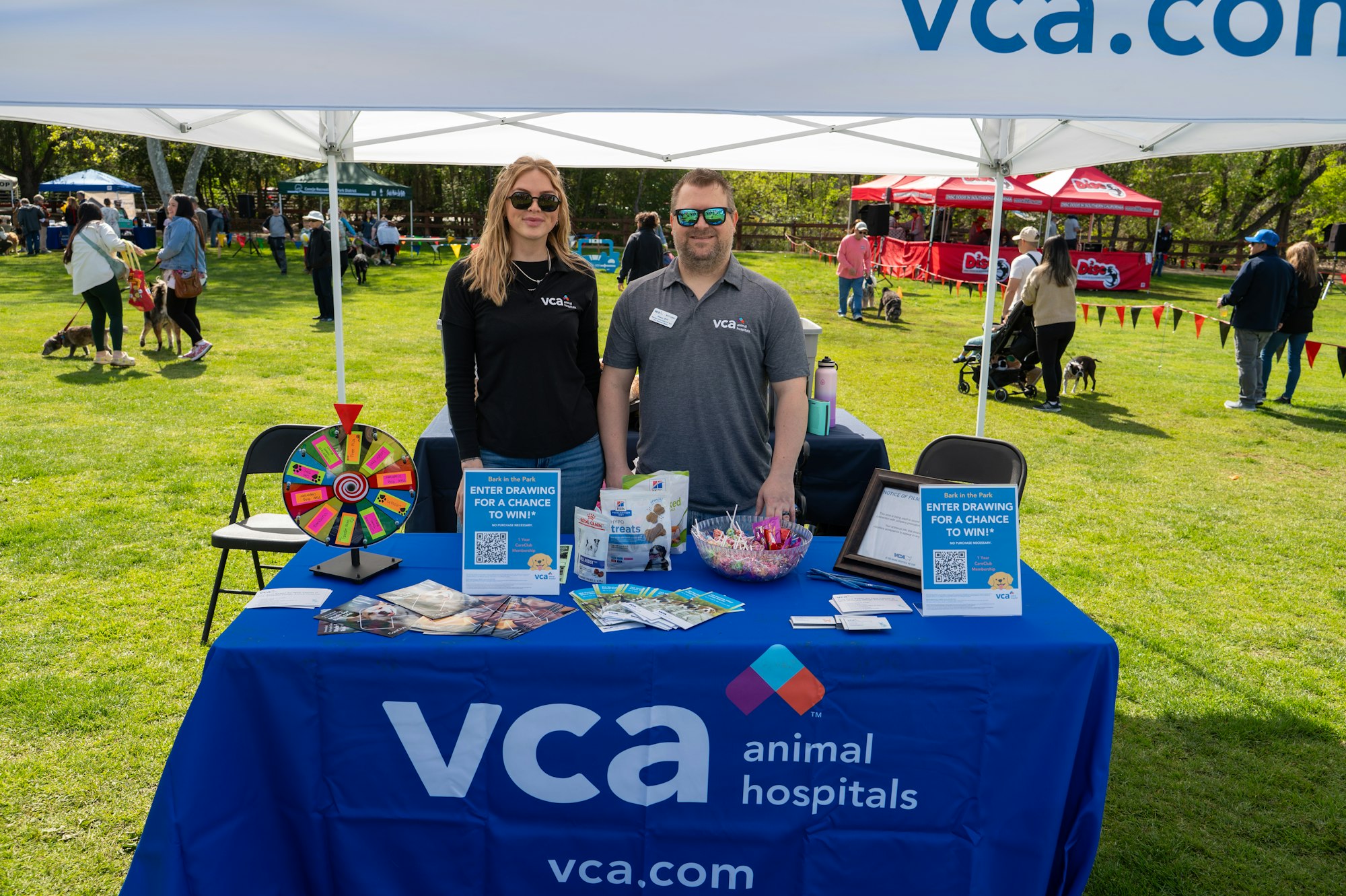 May contain: desk, furniture, table, accessories, glasses, people, person, adult, female, woman, male, man, grass, plant, chair, bag, handbag, clothing, hat, footwear, shoe, animal, canine, dog, mammal, pet, and tent