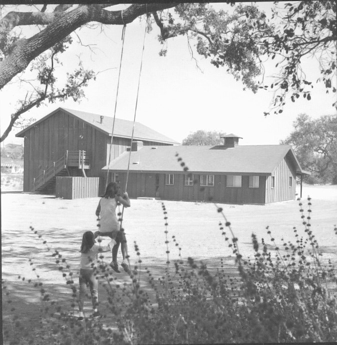 Two kids playing on a wooden swing under a tree near rustic buildings.