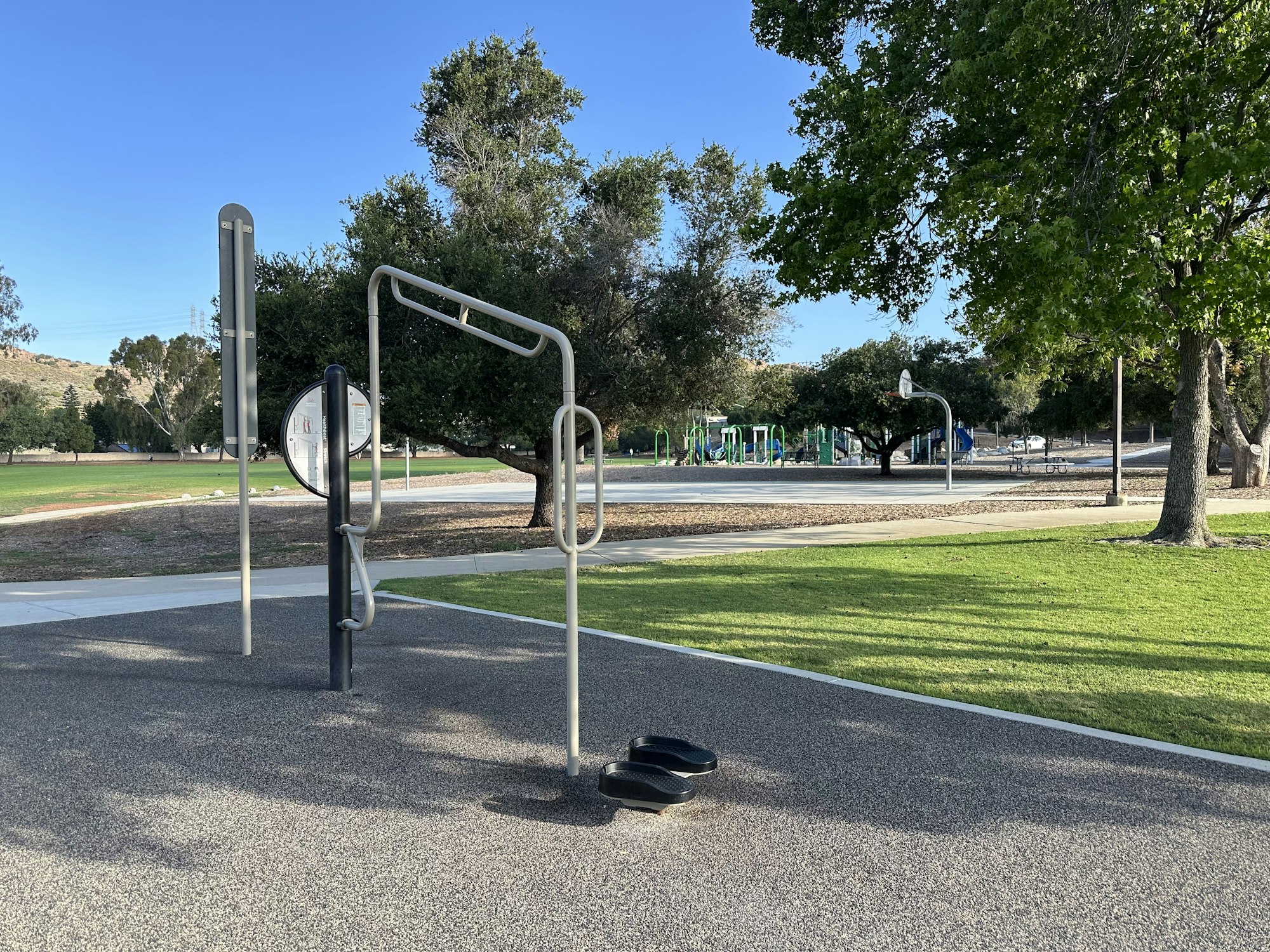 The image shows a park with fitness equipment and a playground in the background, surrounded by trees and green grass.