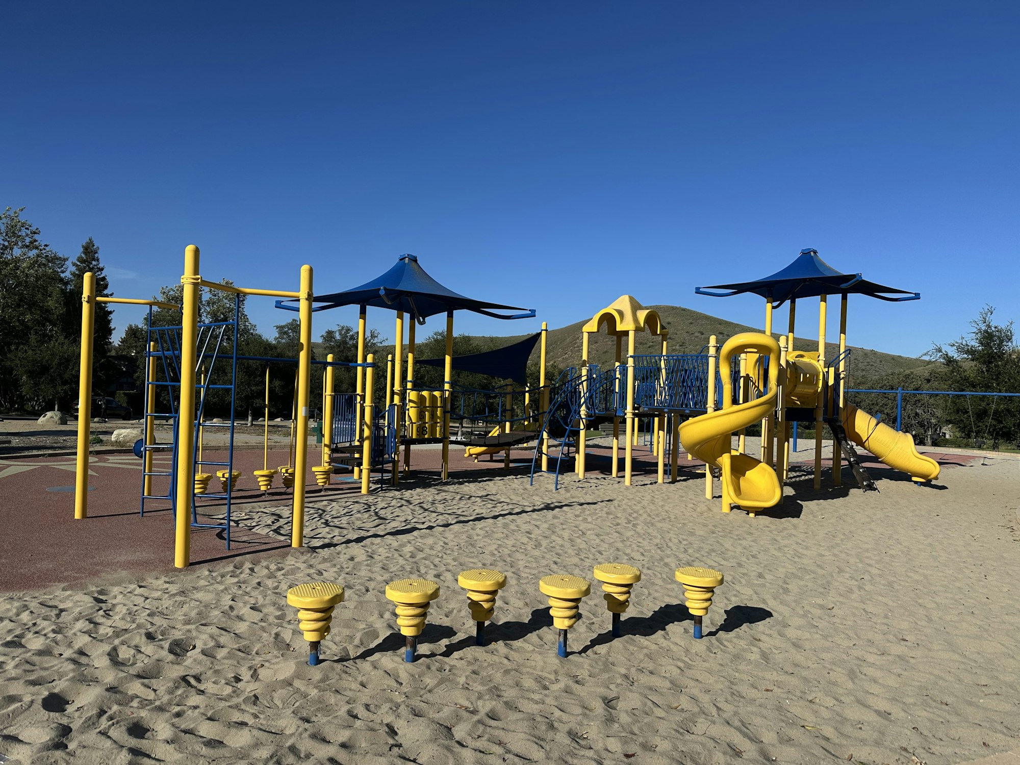 A brightly colored playground with yellow and blue equipment, slides, and climbing structures set on sandy ground under a clear blue sky.