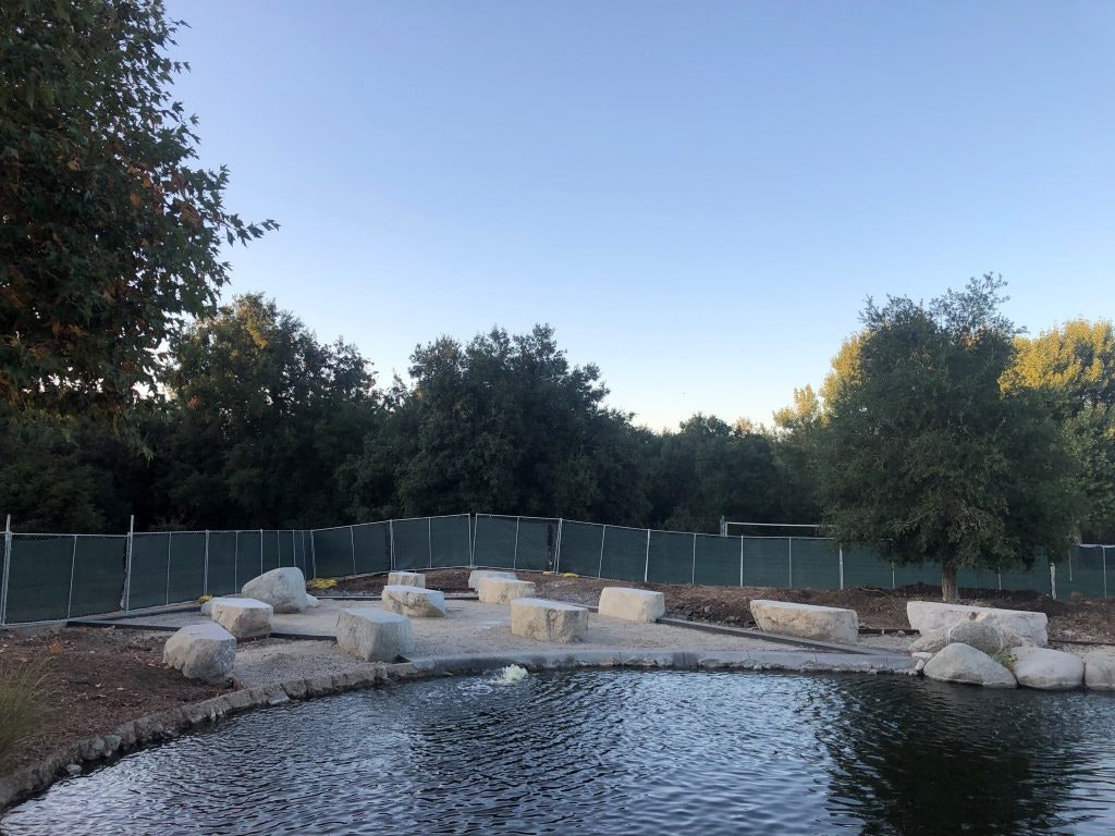 A serene outdoor space with a pond, stone seating, and surrounding trees, partially enclosed by a green fence under a clear sky.