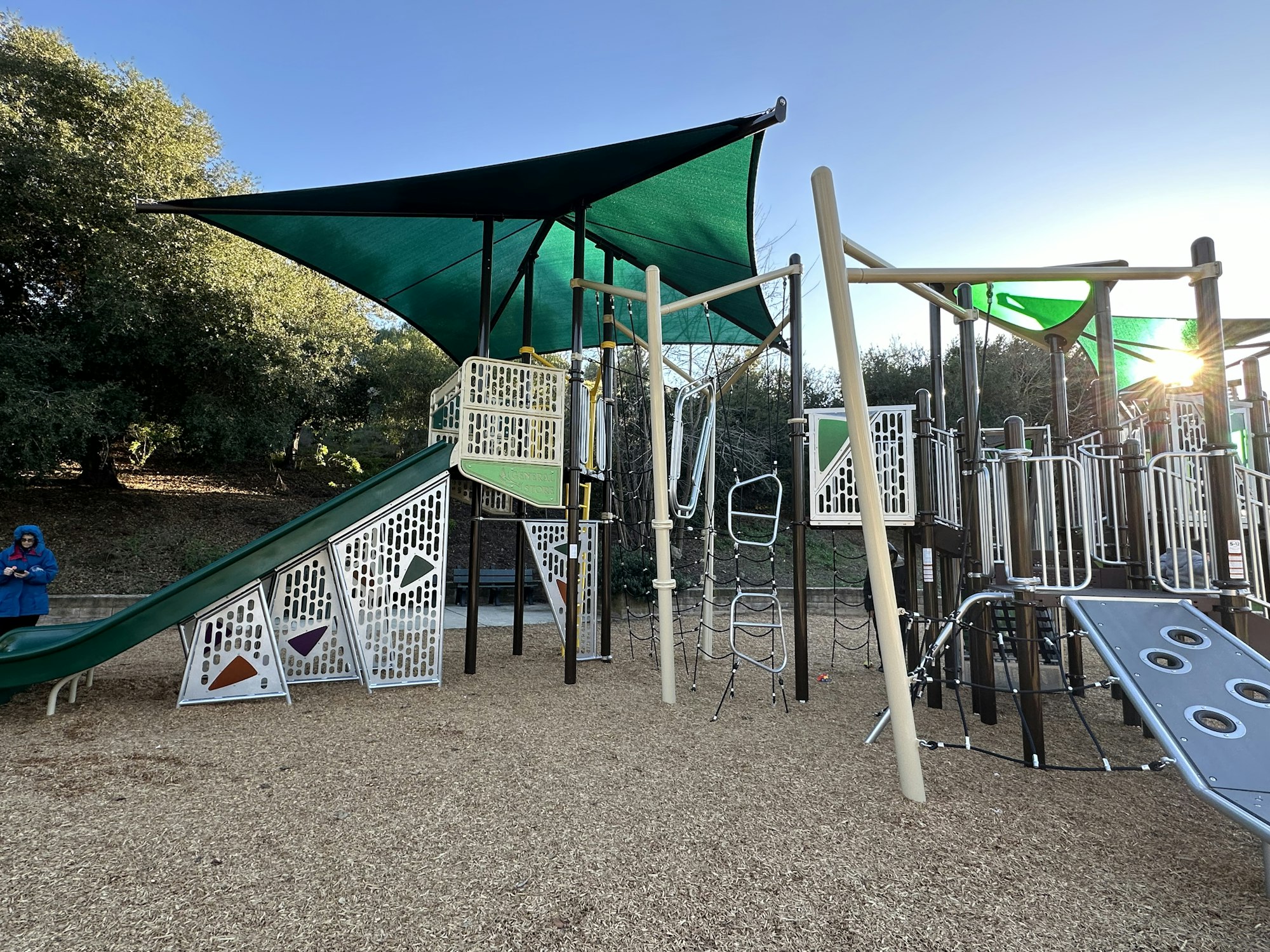 Playground with climbing structures, slides, shade canopies, and a person in a blue coat, surrounded by trees.