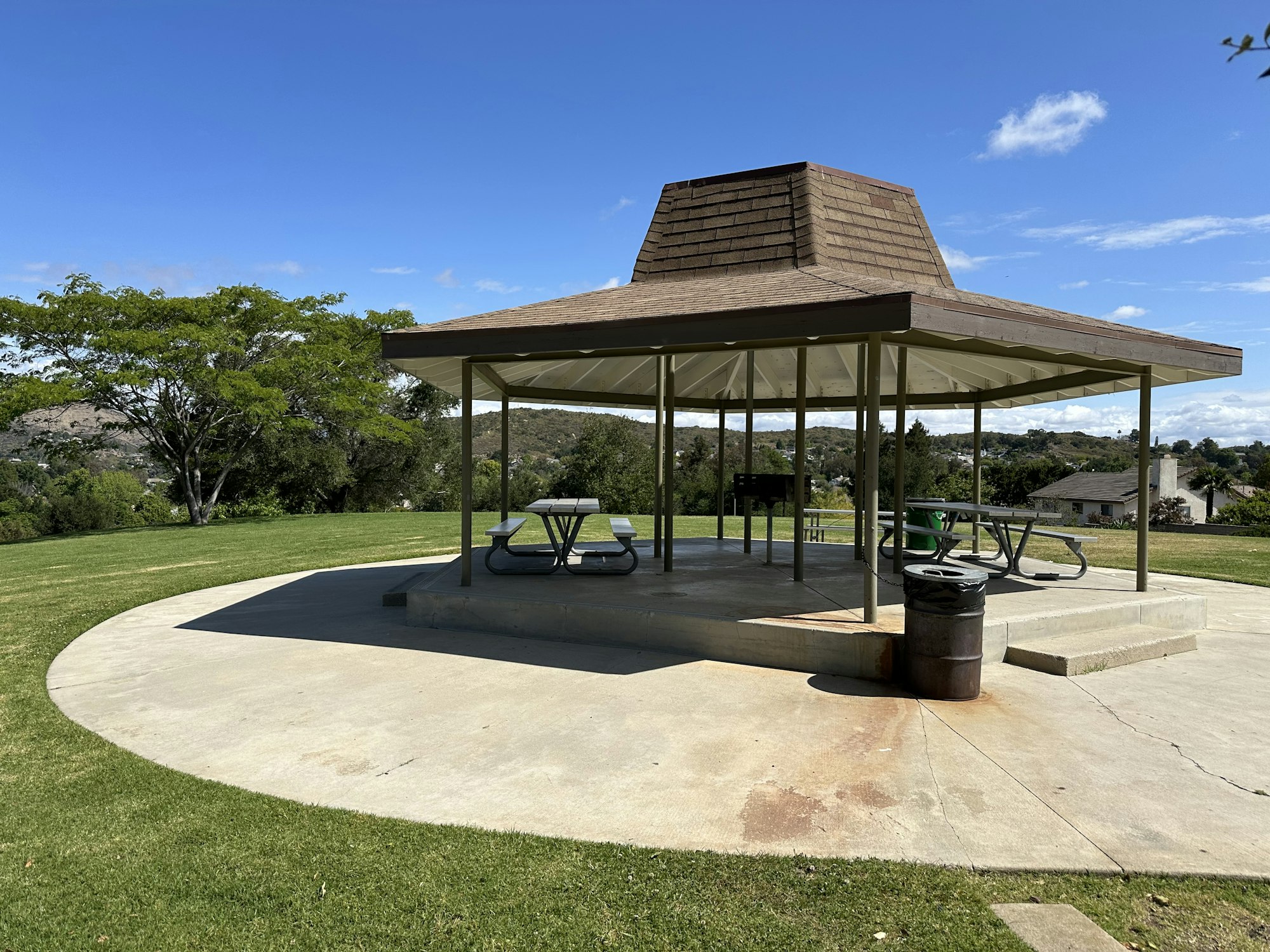 A shaded picnic area with tables, a grill, and a trash can on a grassy lawn under a blue sky.