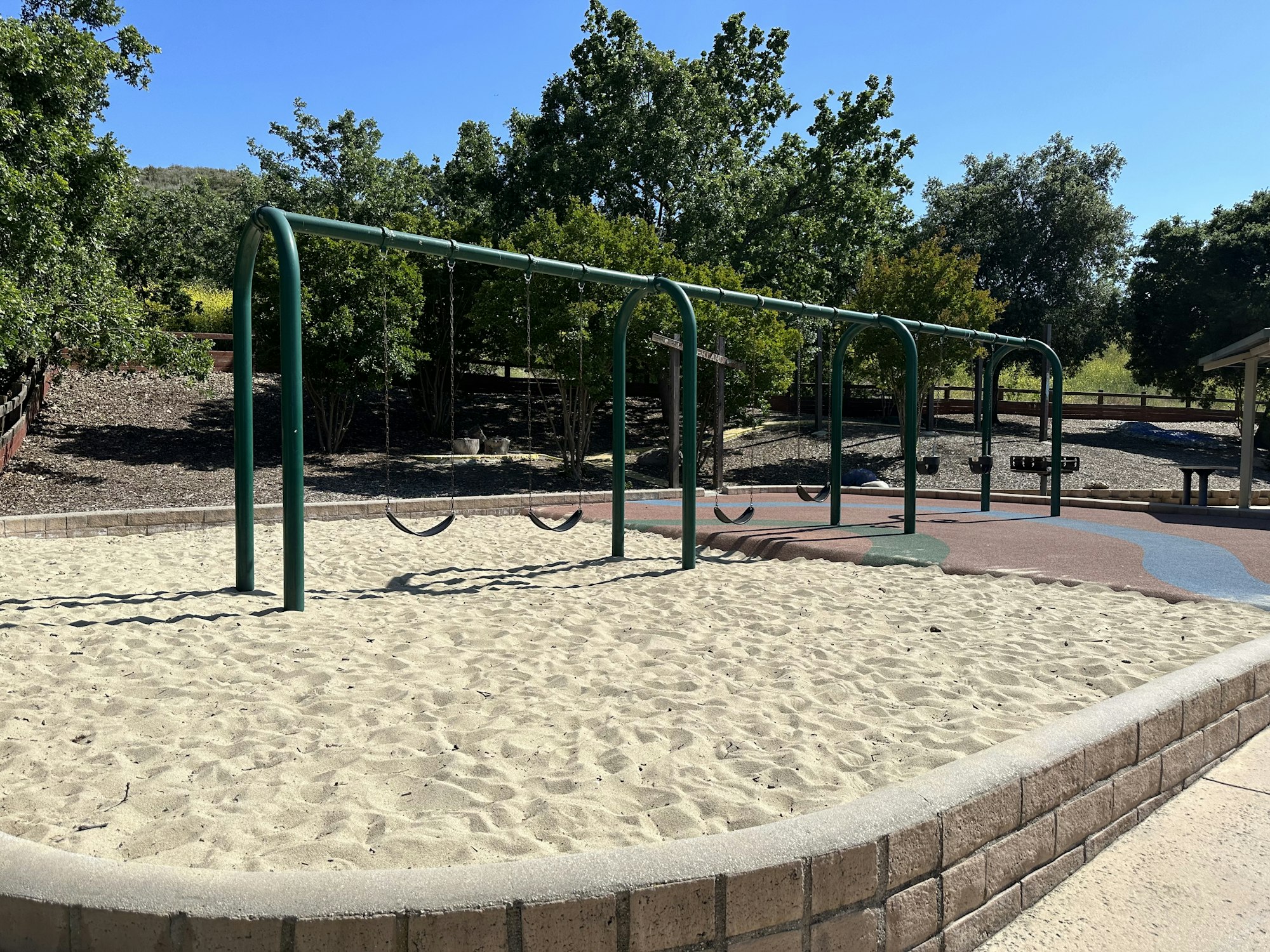 A playground with empty swings on a sandy area, surrounded by trees under a clear blue sky.