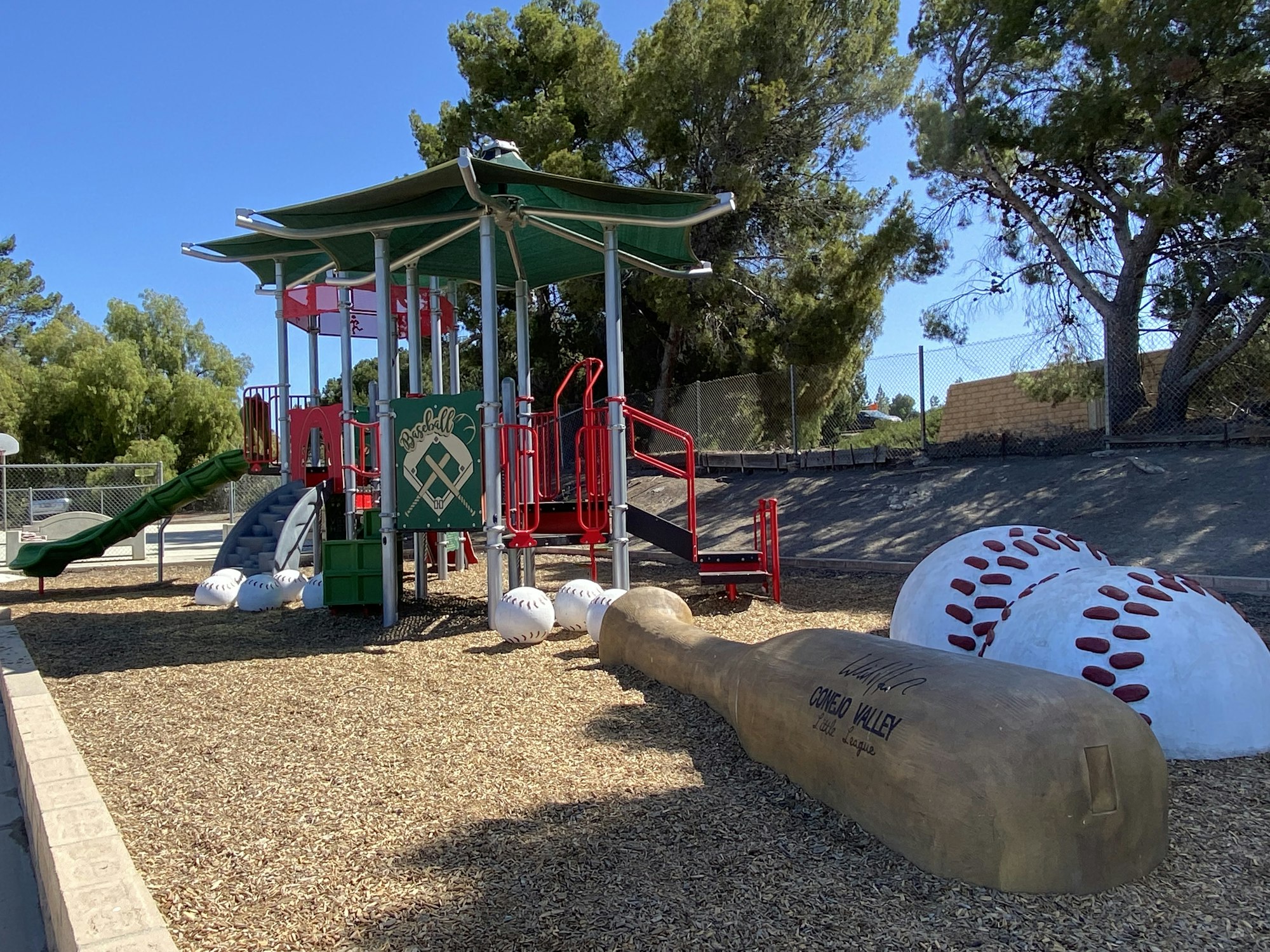 Playground with baseball theme, featuring slides, climbing structures, large baseballs, and a giant bat, surrounded by trees and fencing.